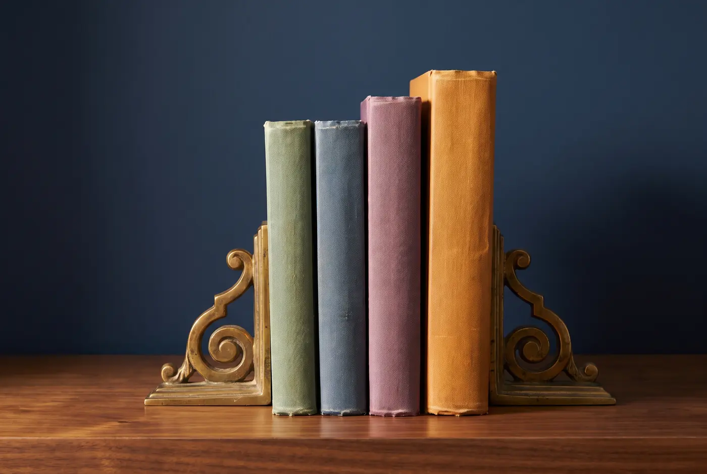 Four cloth-bound ledgers of increasing thickness standing between brass bookends, their spines in sage, slate blue, plum and amber.
