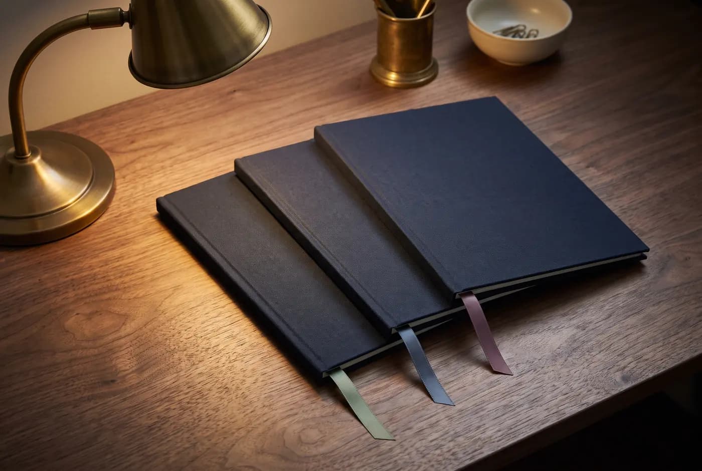 Three navy cloth-bound folders fanned across a walnut desk under a brass lamp, each with a ribbon marker in a different colour.