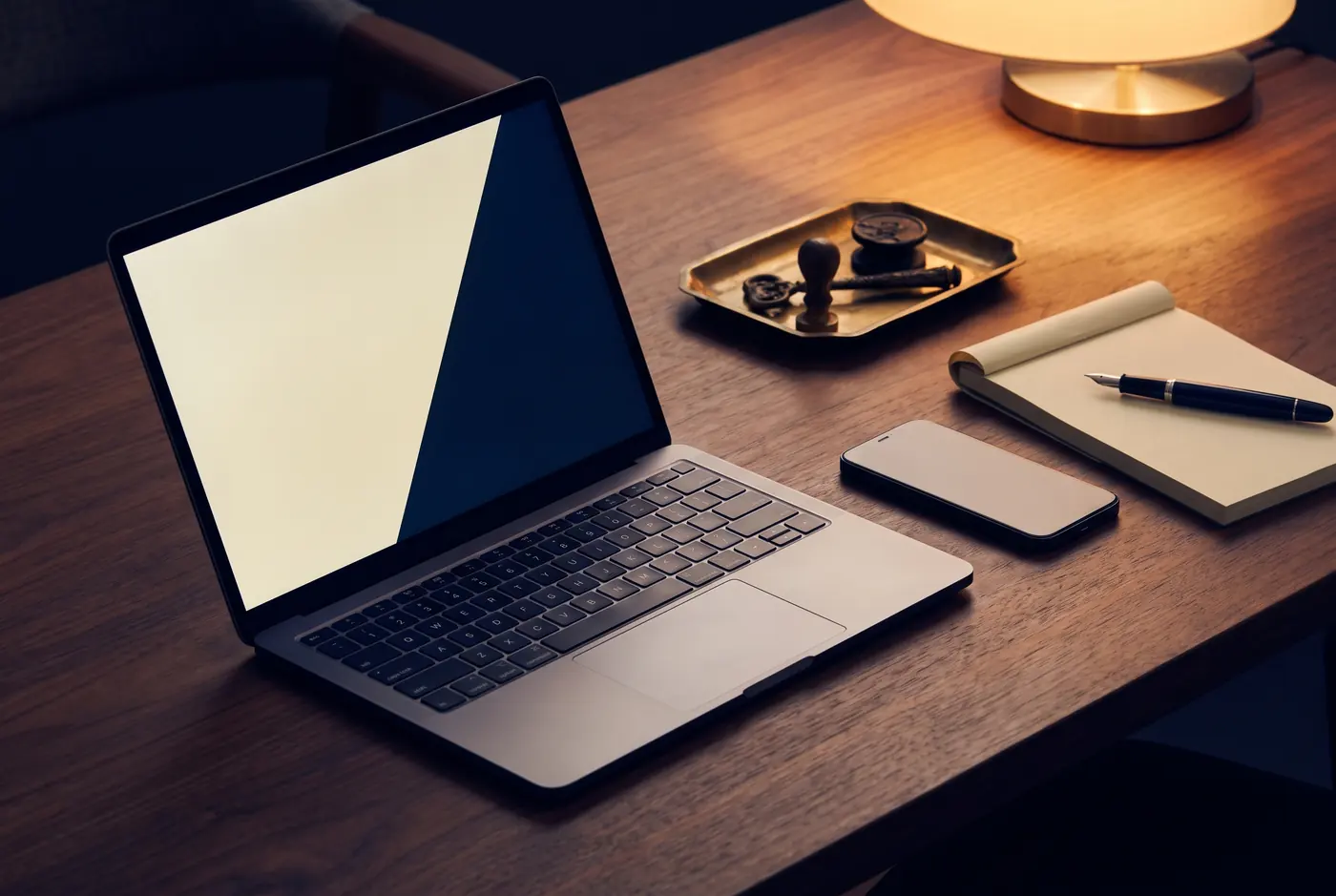 An open laptop on a walnut desk under a warm lamp, its screen showing a plain cream and navy field, with a notepad and a fountain pen beside it.