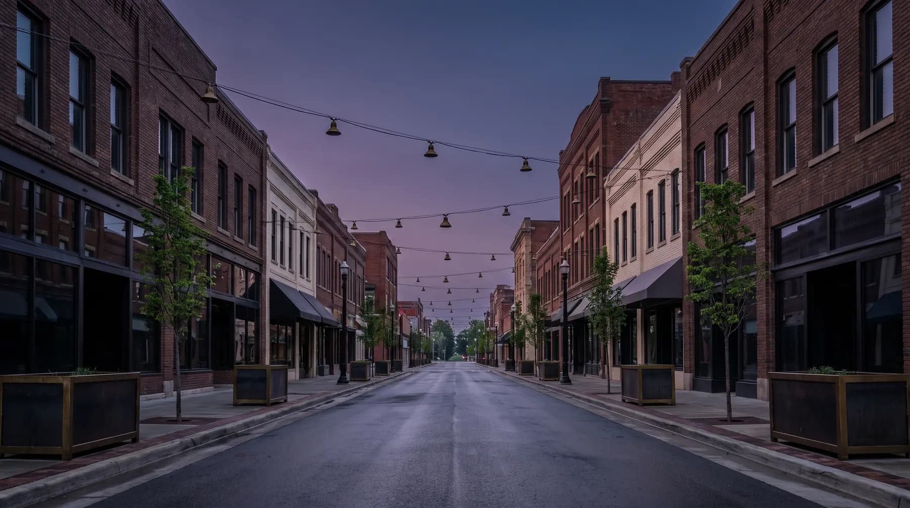 A deserted downtown avenue at dawn, brick buildings with dark windows, unlit string lights strung across an empty street and young trees in planters.
