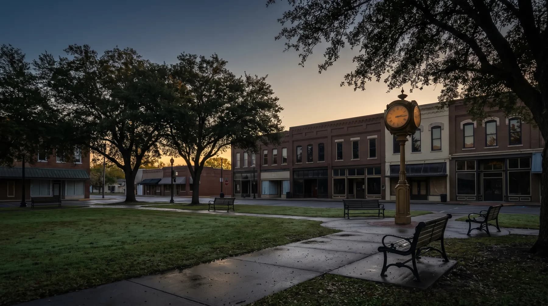A deserted small-town square at dawn: brick storefronts with dark windows around a green with mature oaks, a brass clock tower and empty benches on wet pavement.