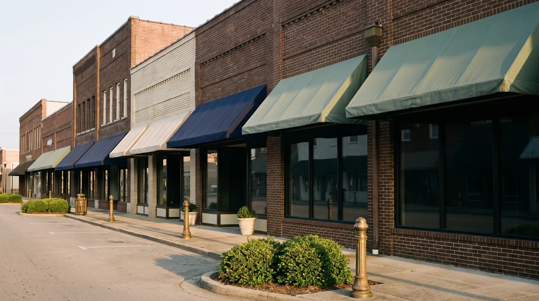 A deserted row of small-town brick storefronts with canvas awnings at dawn, dark windows reflecting the sky, planters of green shrubs on empty pavement.